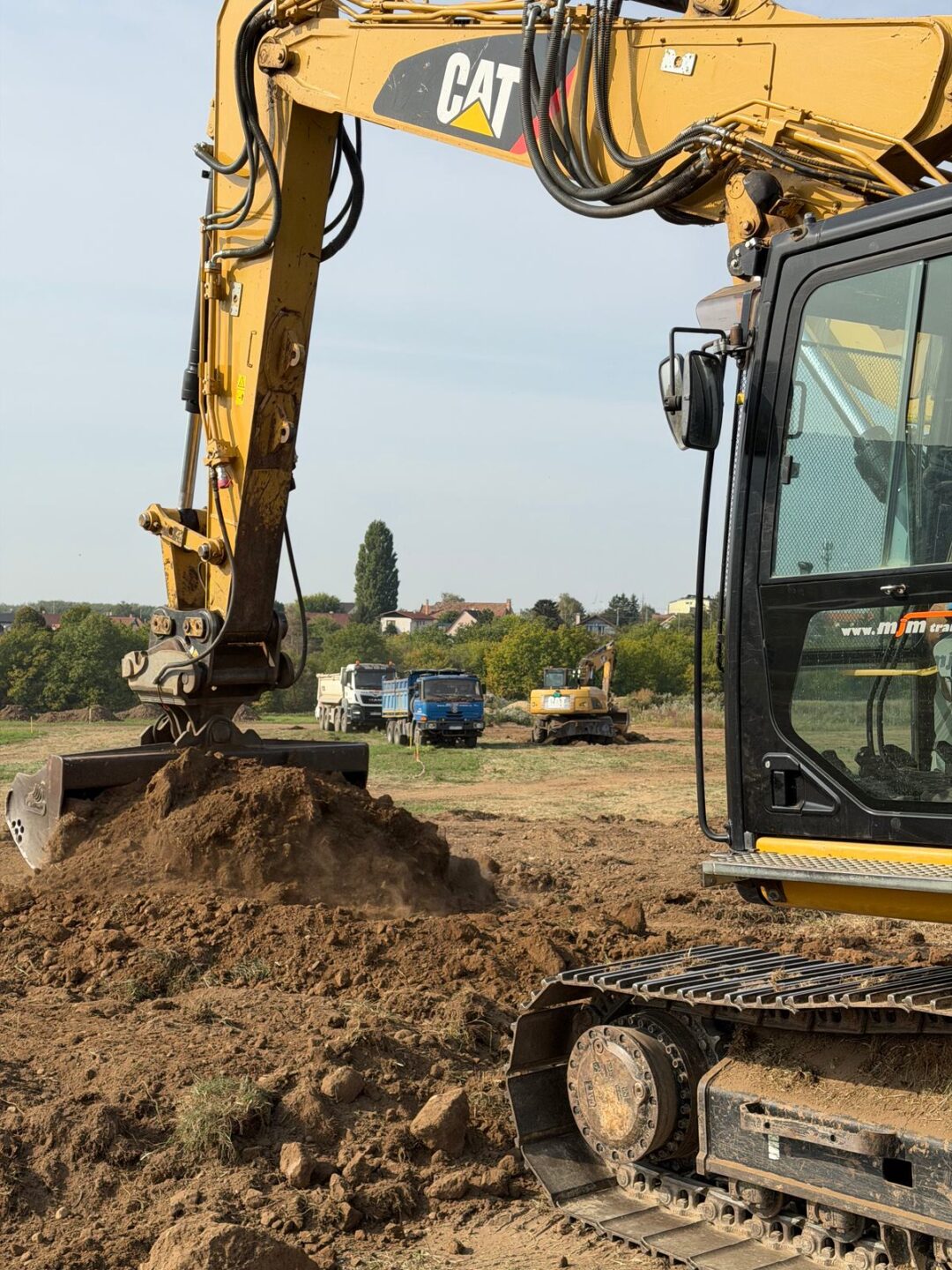 Pásové rýpadlo CAT 320E MJM TRANS pri zemných prácach, v pozadí sklápače