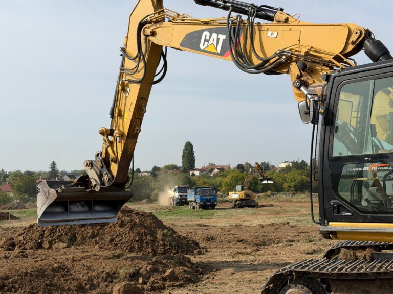 Pásové rýpadlo CAT 320E pri zemných prácach, v pozadí sklápače